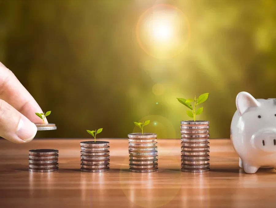 Image courtesy of Healthcare Digital A hand stacking coins next to other stacks of coins. A piggy bank on the right.