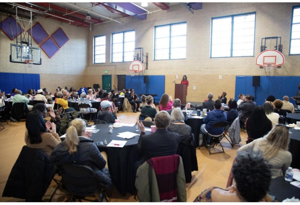 A room of seated people in a gymnasium. A person speaking at a podium at the front.