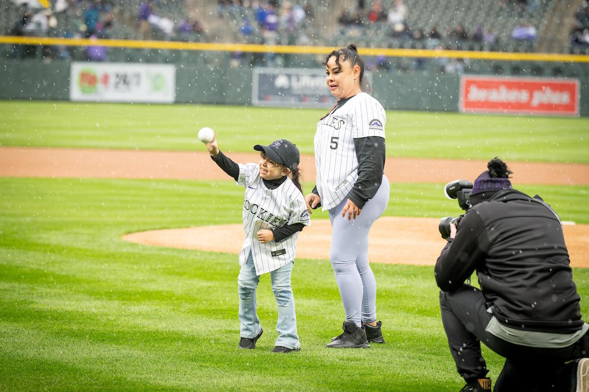 A child throwing a pitch on a baseball field, with an adult behind them and a photographer shown