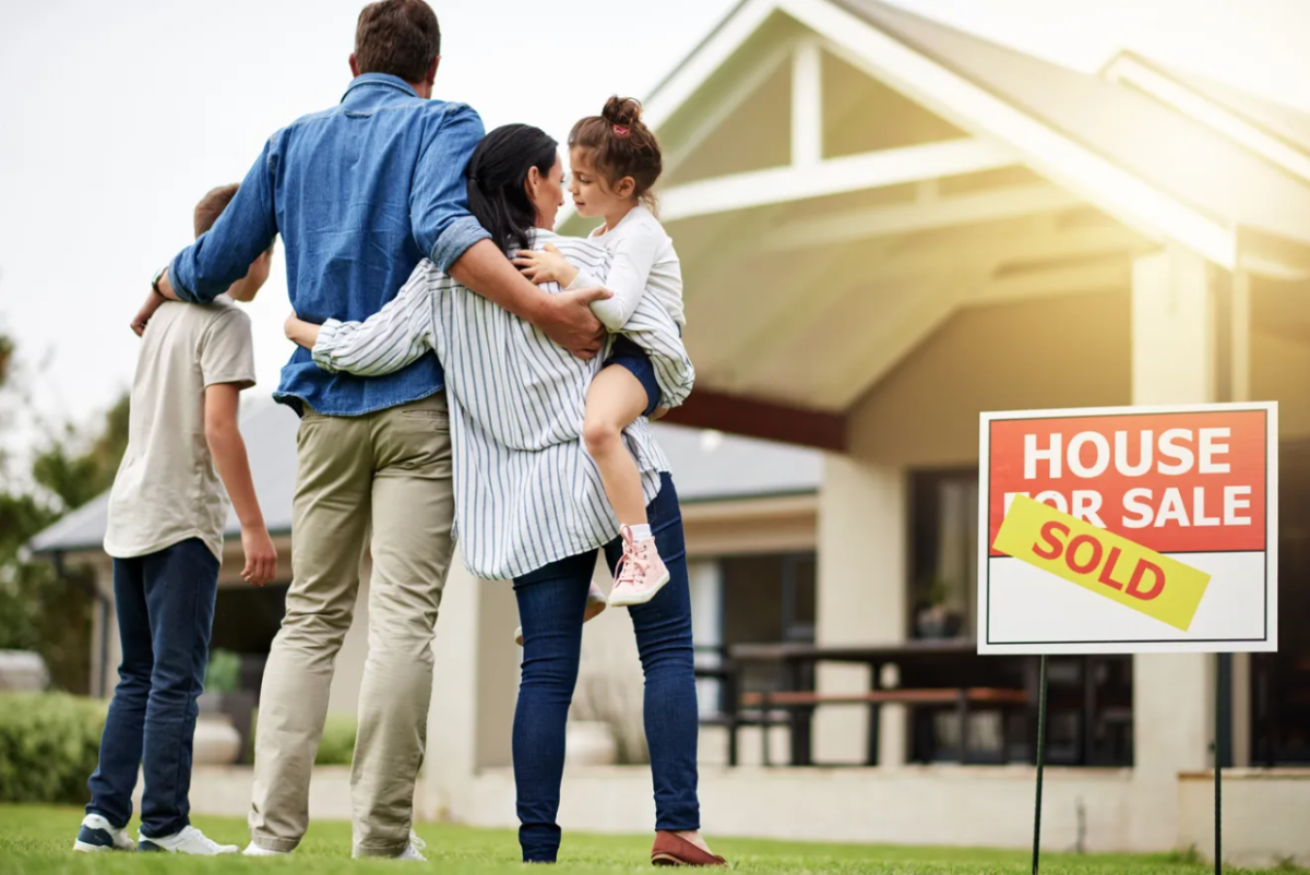 A family standing in front of a hours with a "SOLD" sign in front of it