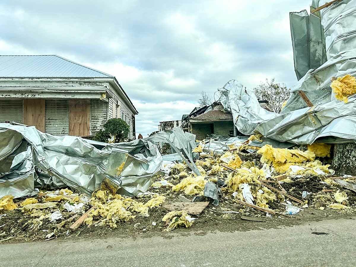 Street view of a tornado-damaged building, insulation strewn everywhere and tarps partially covering it.