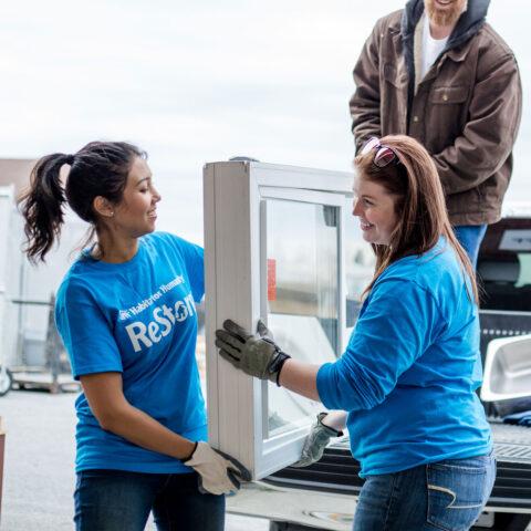 Two people wearing blue tops, working together to carry a large item