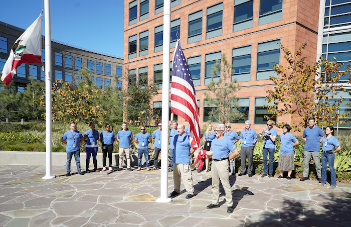Veterans Day 2021 flag-raising ceremony at Illumina headquarters. Two males are raising the flag and a group of Illumina veterans are in a semi-circle around the back of the flag pole. The crowd is made up of 12 men and women.