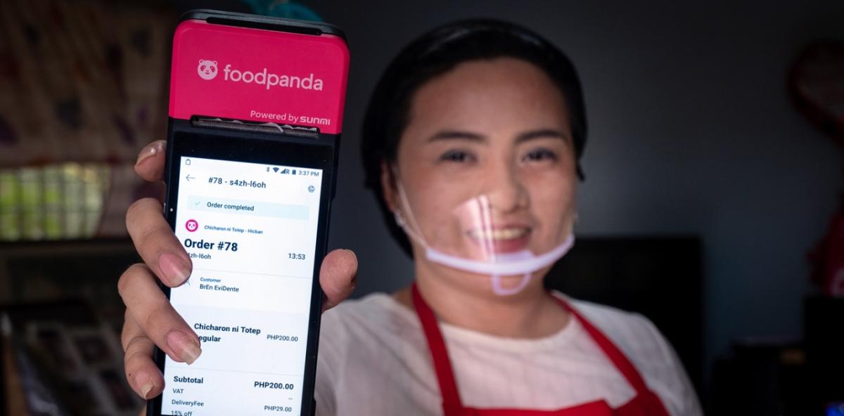 woman in red apron and a plastic face mask holds up a phone featuring the food panda app