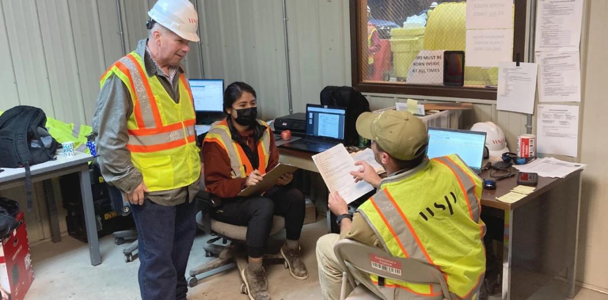 Three workers in an office wearing safety gear, one holds a clipboard and the other a stack of papers