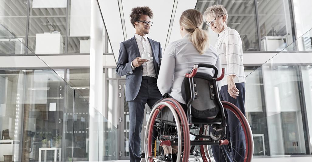 Two people standing and talking with a third in a wheelchair outside a glass-walled building.