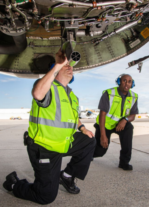 two people kneeling underneath an airplane, both wearing safety vests and hearing protection