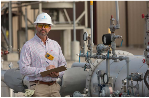 A person in a hard hat writing on a clipboard, next to industrial pipes outside.