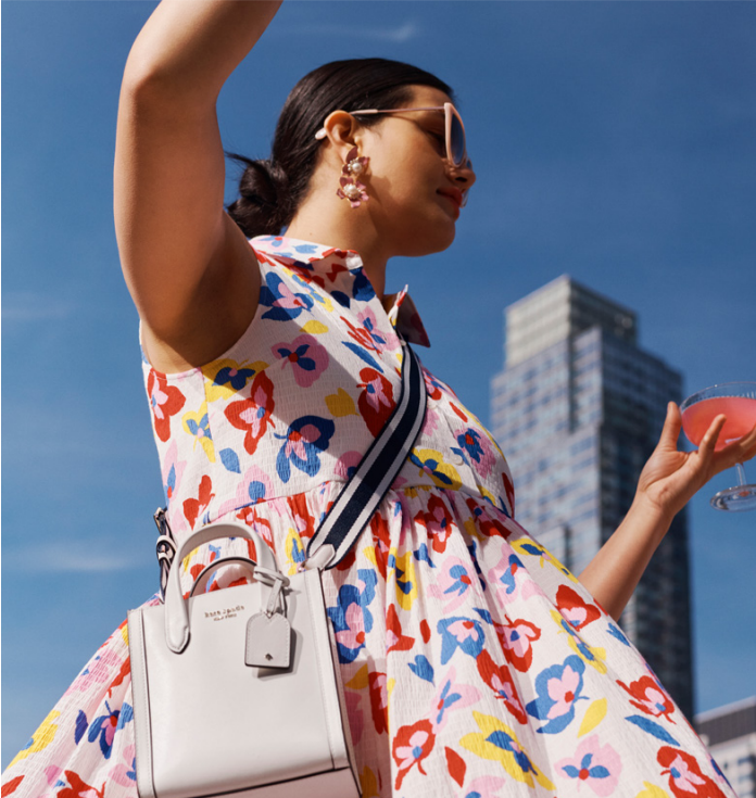 A person in a colorful kate spade dress, sunglasses, and cross-body handbag. A tall building behind them. 