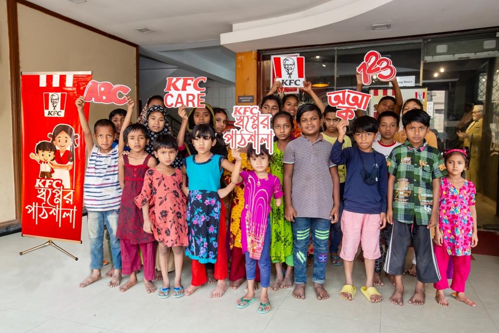 A group of standing children, holding signs of the KFC logo, KFC Care, ABC, 123, and those in another language.