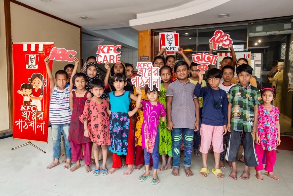 Image courtesy of Dhaka Tribune A group of standing children, holding signs of the KFC logo, KFC Care, ABC, 123, and those in another language.