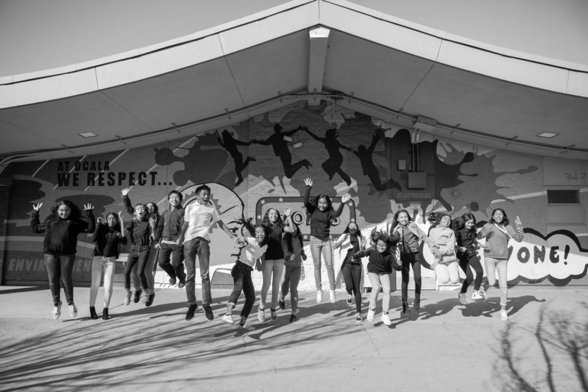 Black and white photo of a group of kids in a line jumping. A covered stage behind them.