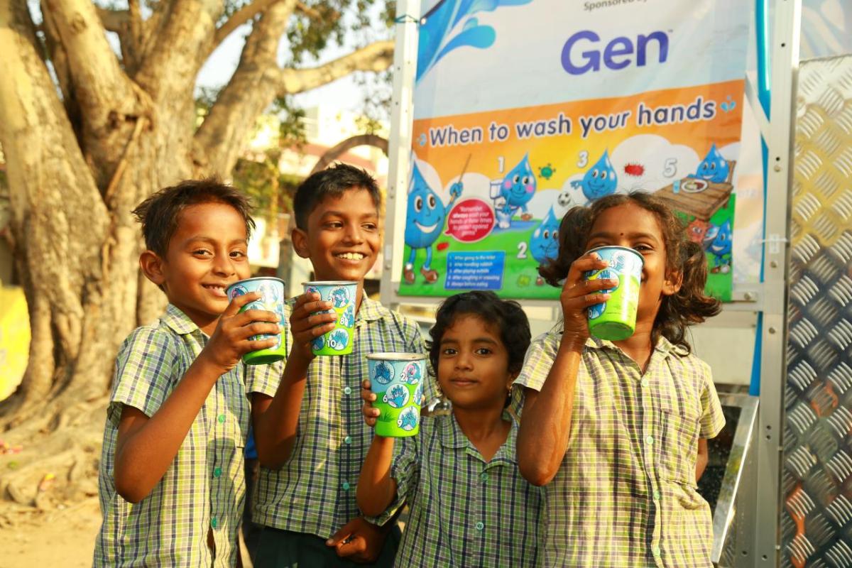 A group of smiling children holding out cups. A sign "Gen. When to wash your hands" behind them.