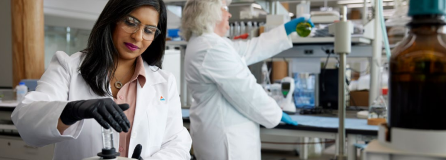 Two people in a laboratory setting, wearing white lab coats and protective gloves and eye wear. 