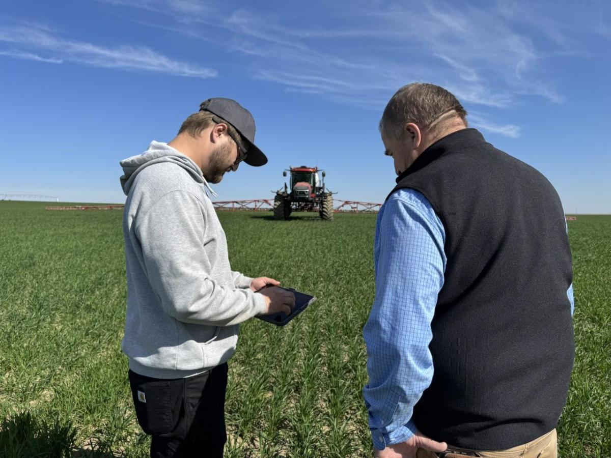 Two people standing together in a field looking at a screen, with a sprayer in the background