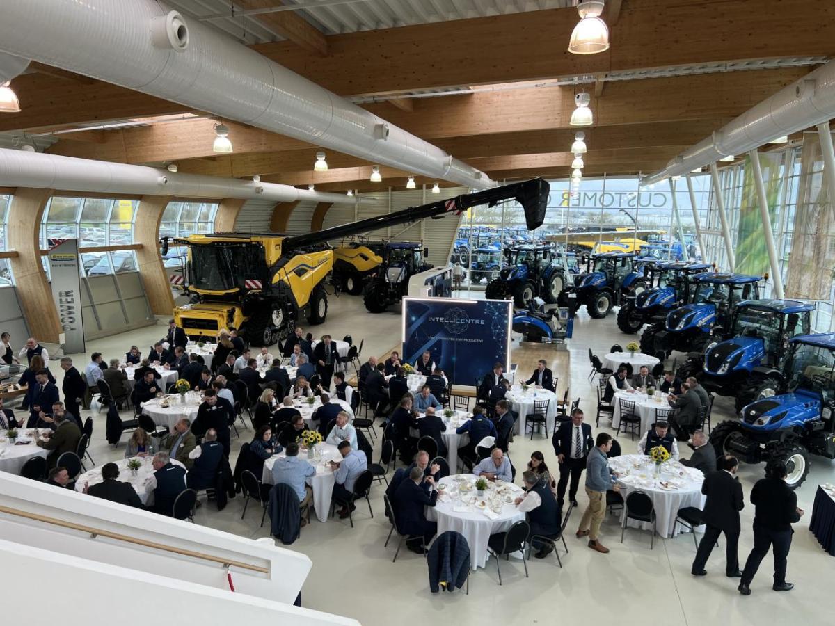 Wide view of a group of people seated at circular tables. Large farming and construction vehicles behind them.