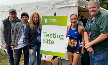 a group of five people, one in running apparel, by a sign for Quest "Testing Site" and a white tent behind them