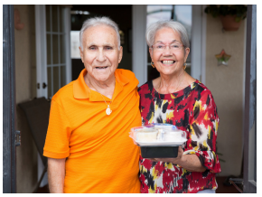Two elderly people smiling on a porch, one holding a container of food.