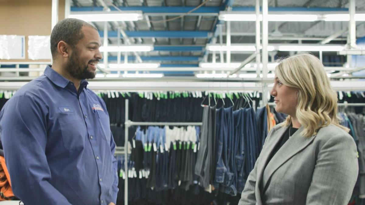 Two people talking, rows of hanging clothes behind them.