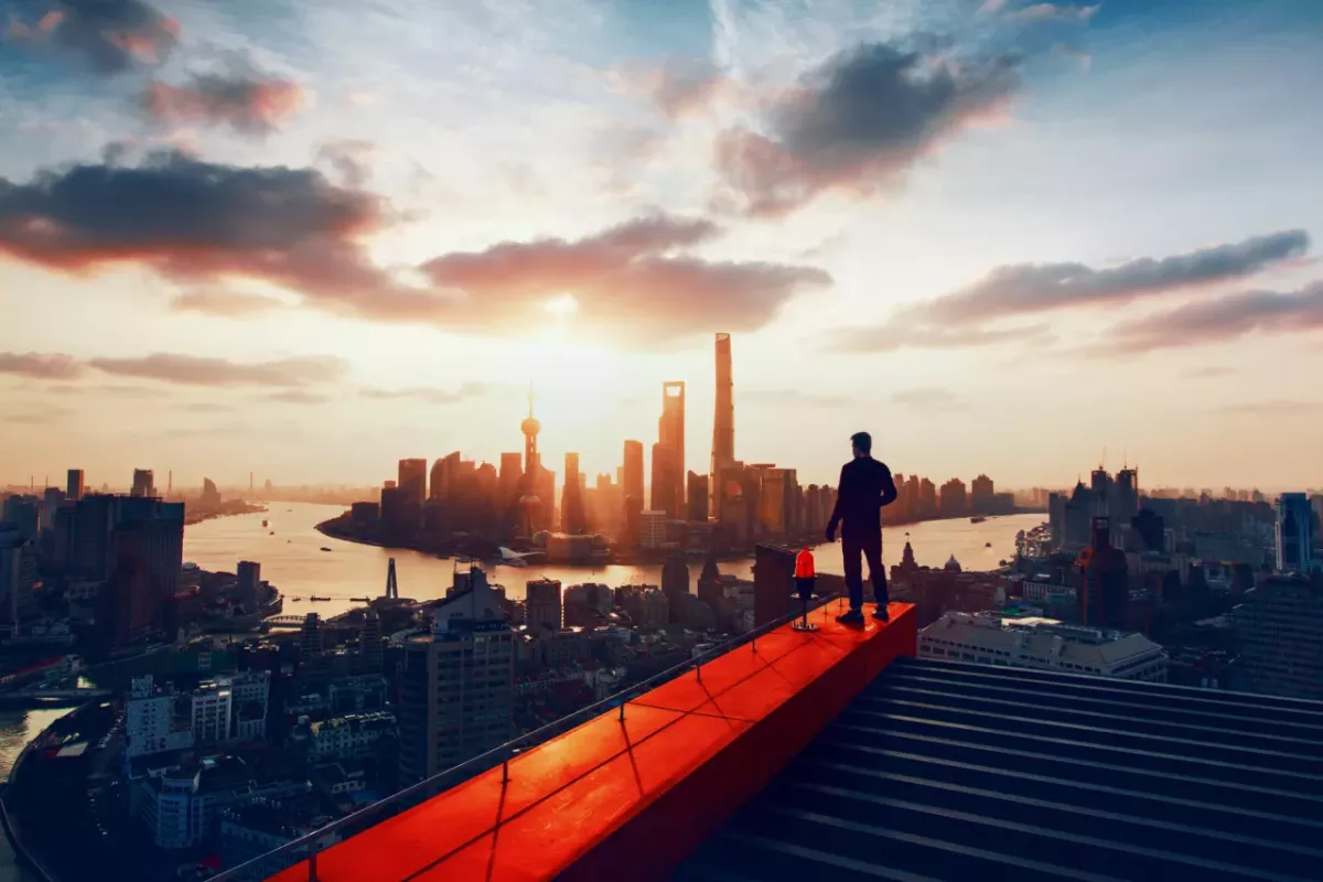 A person standing on a construction beam on the top of a very tall building, overlooking a city skyline and river.