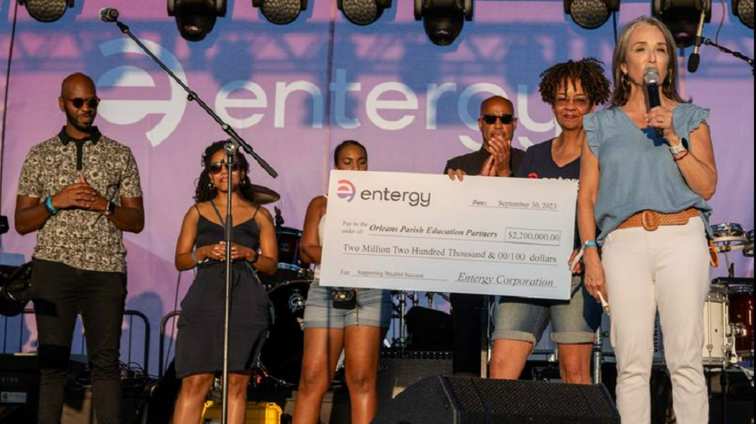 A group of people on a stage. One holds a microphone as another behind them holds a large check.