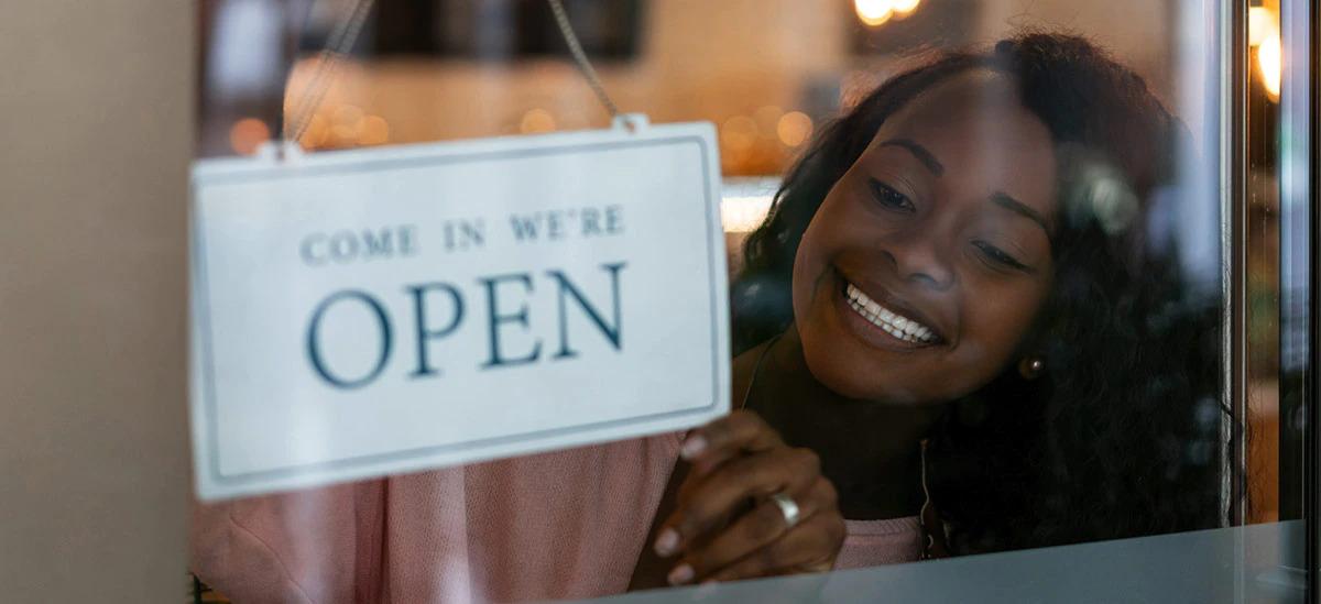 A smiling person flipping a sign to say "Come in we're OPEN" 