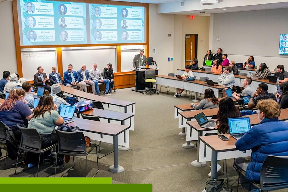 A speaker at a podium in a room of seated others. A digital display behind them with profiles of the panelists.