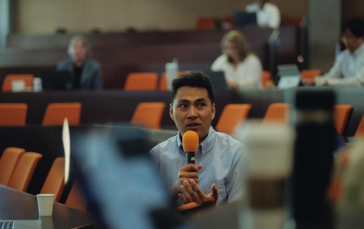 A person seated in an auditorium talking into an orange mic.