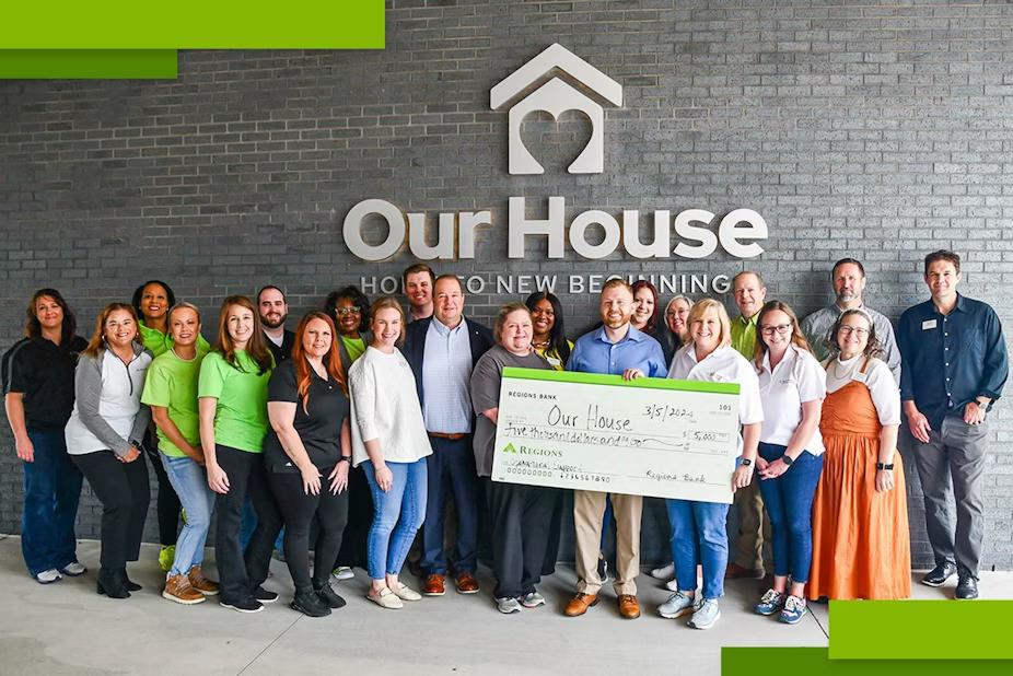 A greoup of people posed in front of the side of a building "Our House" sign above them. Some holding a large check made to "Our House".
