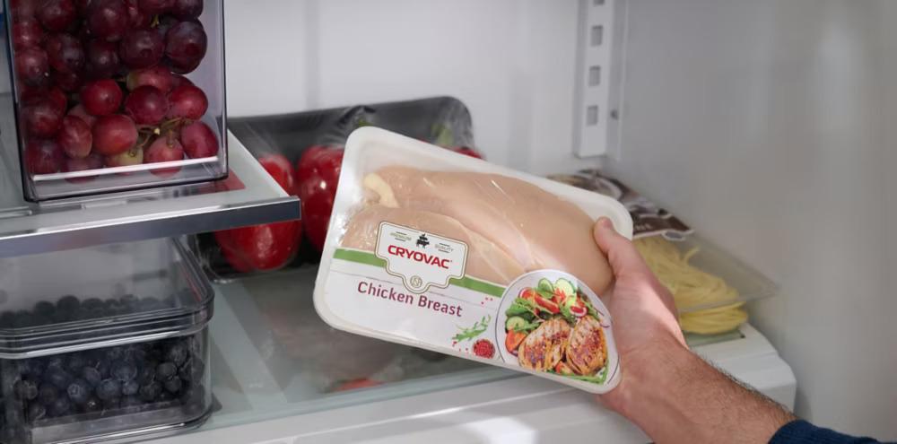 A hand holding a package of "Chicken Breast" in a refrigerator.
