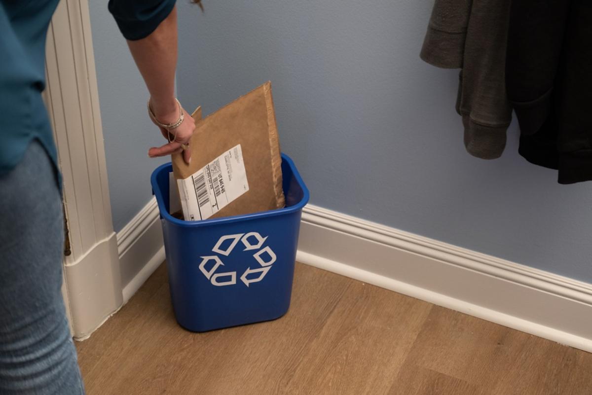 Closeup of a person's hand putting a paper packaging mailer into a small recycling bin inside a home.(