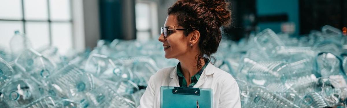 A person in a lab coat with a clip board in front of a large pile of clear plastic containers.