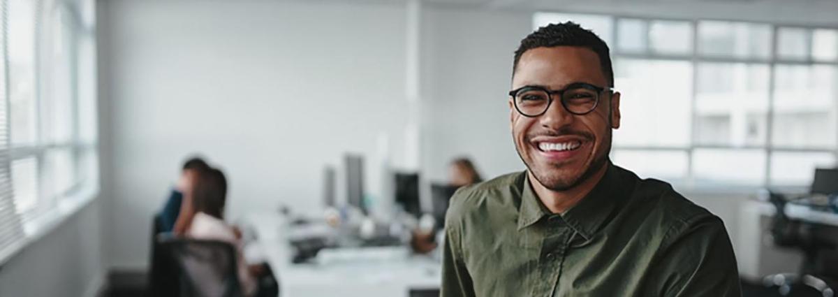 A smiling person looking at the camera, a small group of people at a shared desk behind them, in an office setting