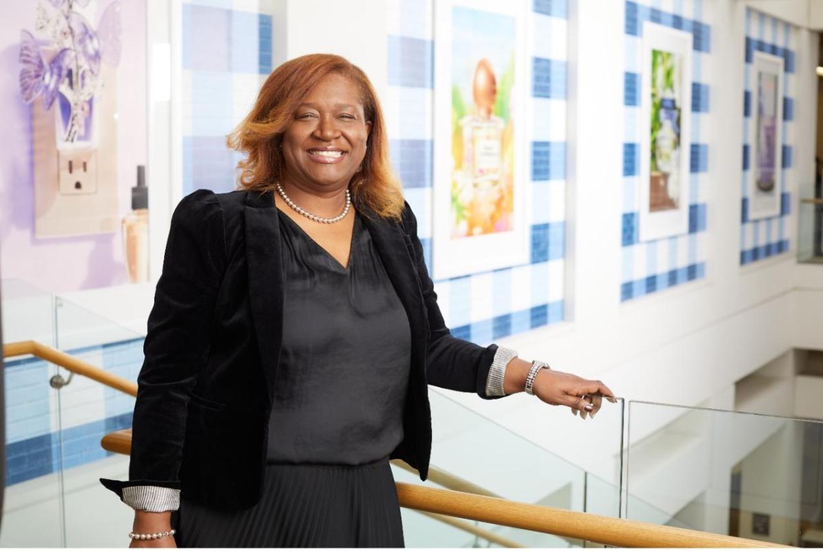 Kelie Charles is pictured in a black suit smiling at the camera with images of Bath & Body Works products are displayed on a wall behind her.