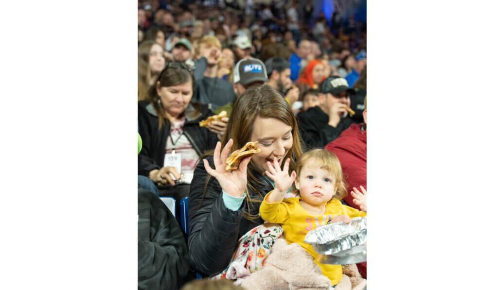 A crowd of people in the stands, close up of an adult and child waving, a piece of pizza in their hands.
