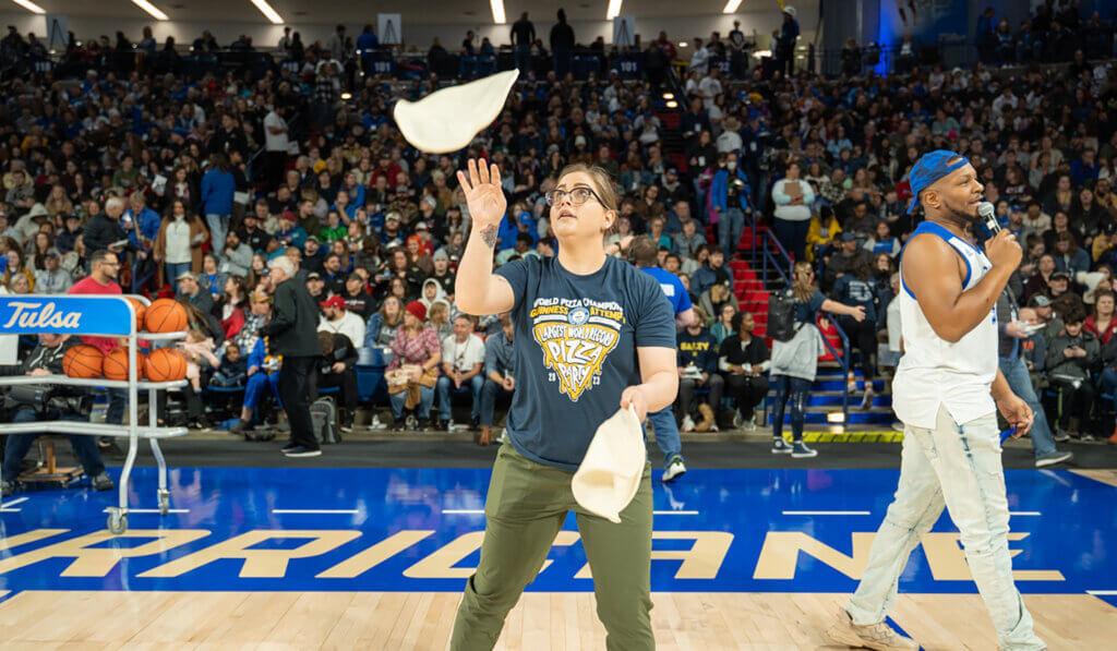 A person standing on a basketball court tossing two pizza dough crusts. A person walking behind them with a microphone. A large crowd in the stands.