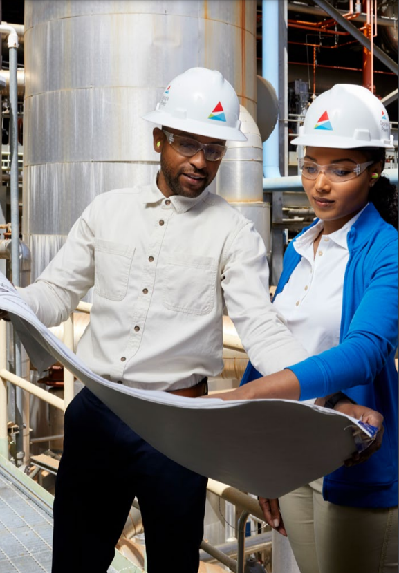 Two people in business casual attire, hard hats and protective eye ware looking over a blueprint. They are standing in a large plant.