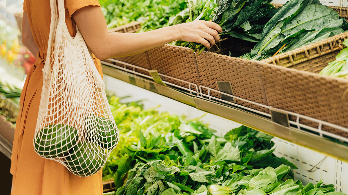 shopper carrying a mesh bag selecting leafy greens from wicker baskets