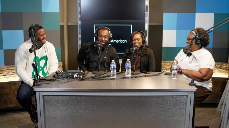 Four people at a large table, each wearing headphones and have a microphone in front of them.