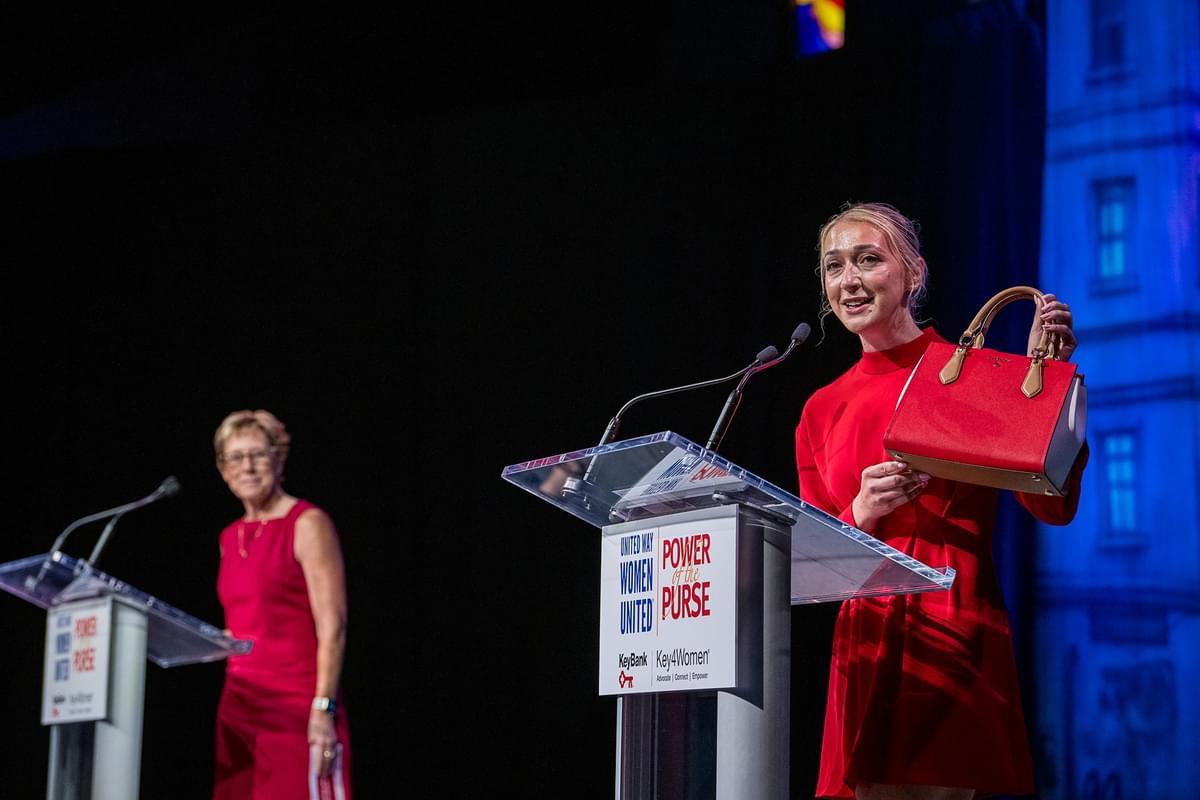 person standing at event podium holding a red purse