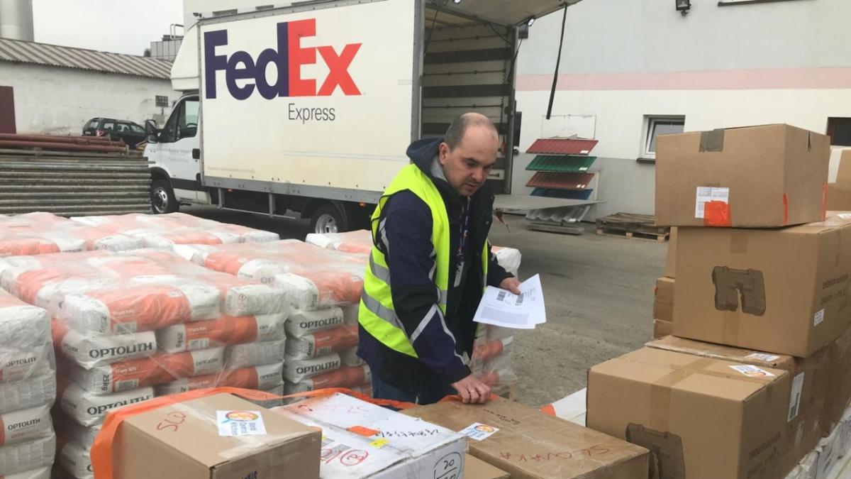 a person checks rows of boxes and wrapped pallets. A fedex truck behind the.