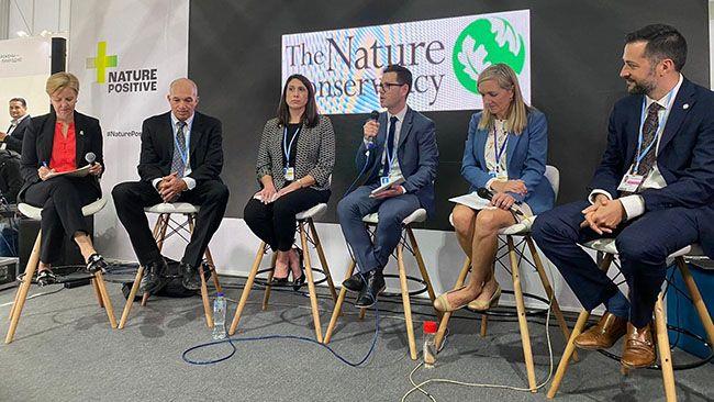 Six people seated on tall chairs. Middle person speaking with a microphone. All in business attire. A banner "The Nature Conservancy" behind them.