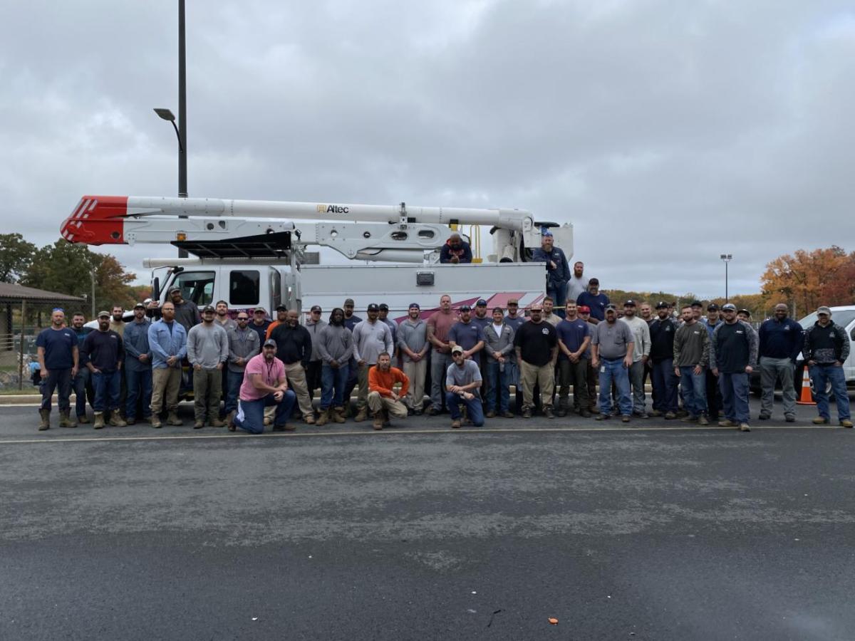 A large group of people posed outside in a parking lot in front of a truck with large extension arm and bucket.