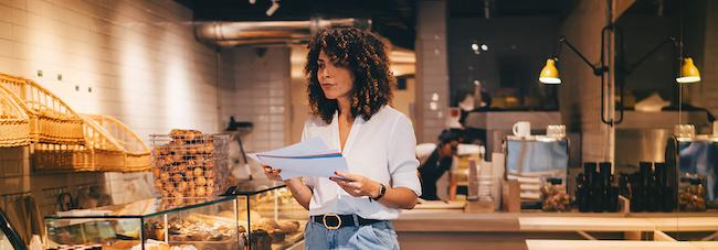 Woman standing in a restaurant, looking over paperwork. Restaurant is a bakery/coffee shop.