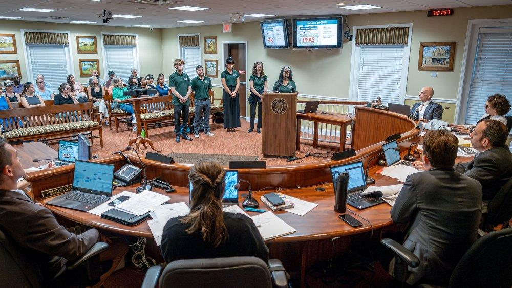 Each summer, the PSEG Institute for Sustainability Studies Green Teams program helps community and corporate partners solve environmental and climate challenges. Shown, 2024 student interns present their solutions to environmental and health risks and benefits of grass versus artificial turf to city officials in Verona. (Photo by Mike Peters) Five people standing together in the center of a room, surrounded by people sitting with laptops