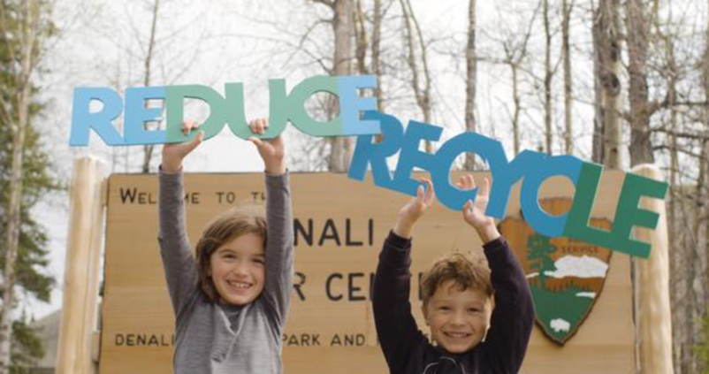 Two children stand in front of a Denali park sign holding up words "Reduce" and "Recycle"
