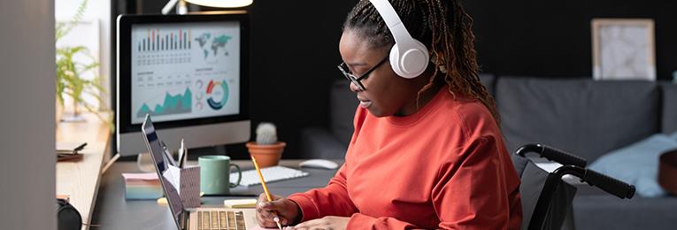 A person with headphones working at a desk by a window with a pencil in hand and an open laptop and separate screen.