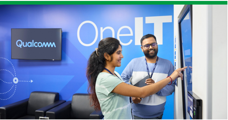 Two smiling people looking at a large touch-screen display. "OneIT" on the wall behind them.