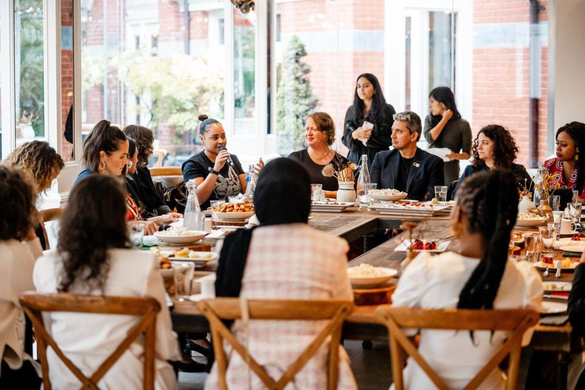 People seated at a dining table. One speaking with a microphone.