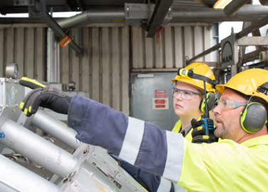 Two people in safety wear in front of industrial piping. One pointing to a gauge.